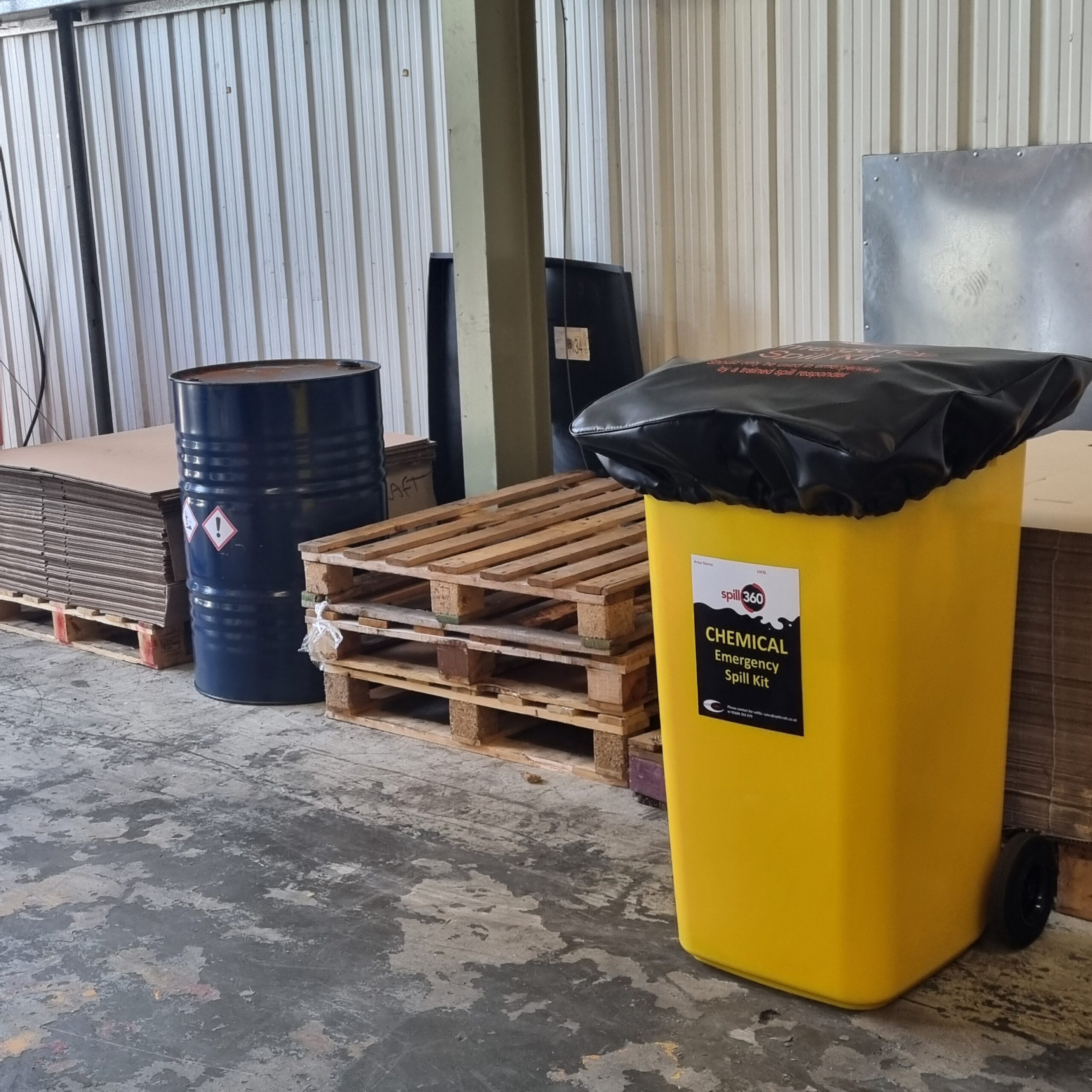 A yellow chemical emergency spill kit bin standing next to wooden pallets and a blue oil drum in an industrial warehouse.
